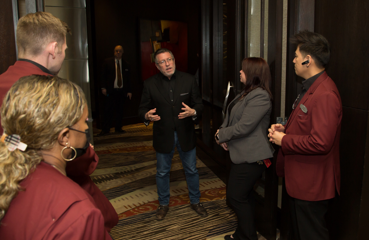 A man in a black jacket speaks to a group of people in uniforms in a hallway at the National Conference Center, fostering discussion among the Braintrust.