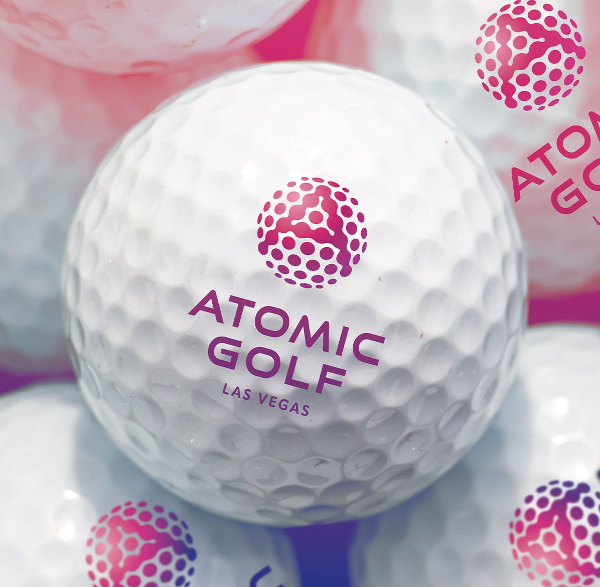 Close-up of white golf balls with Atomic Golf Las Vegas logo in pink and purple lettering.