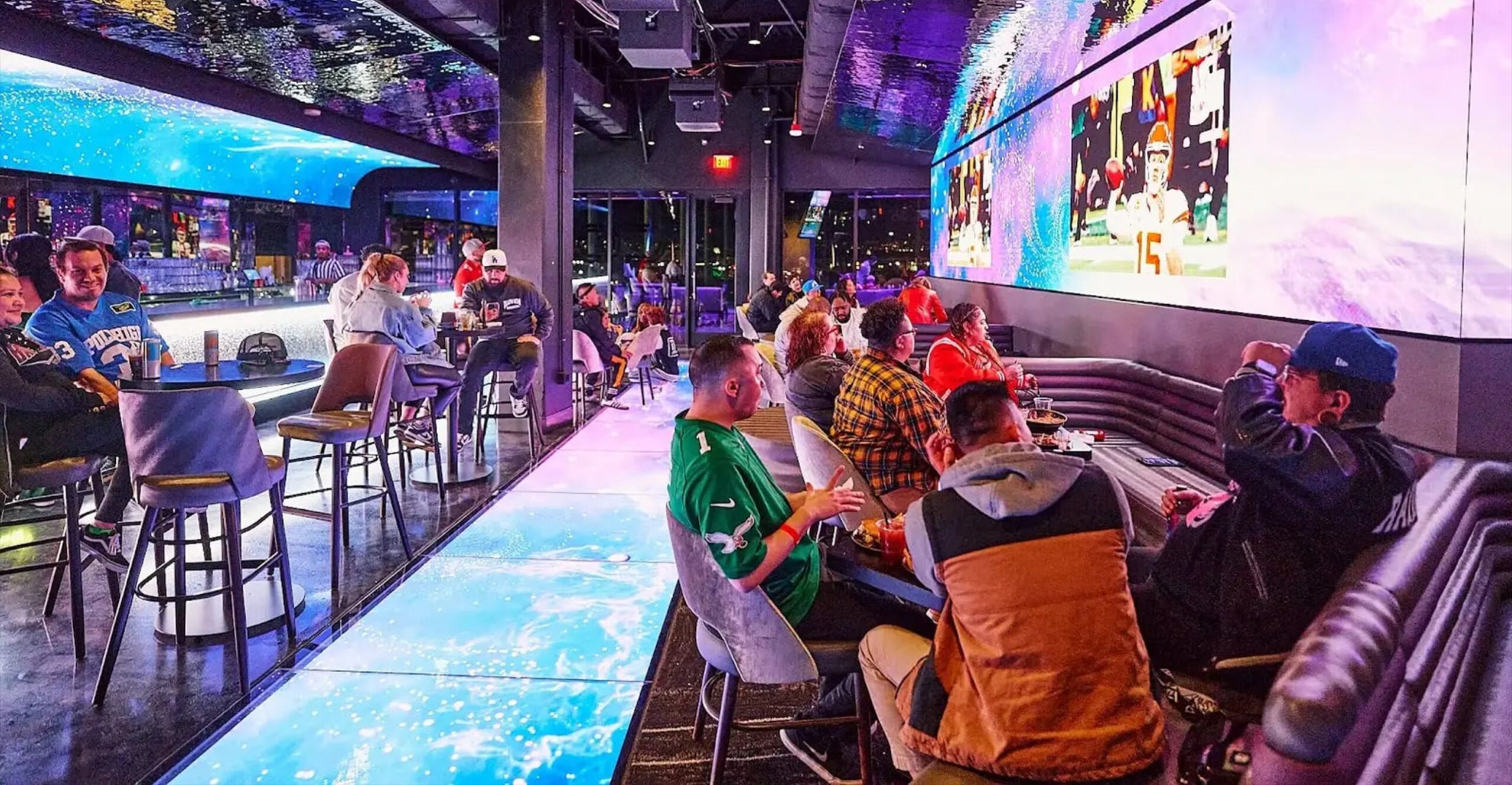 People sit at tables in a lively sports bar with big screens, neon lights, and a glowing floor.