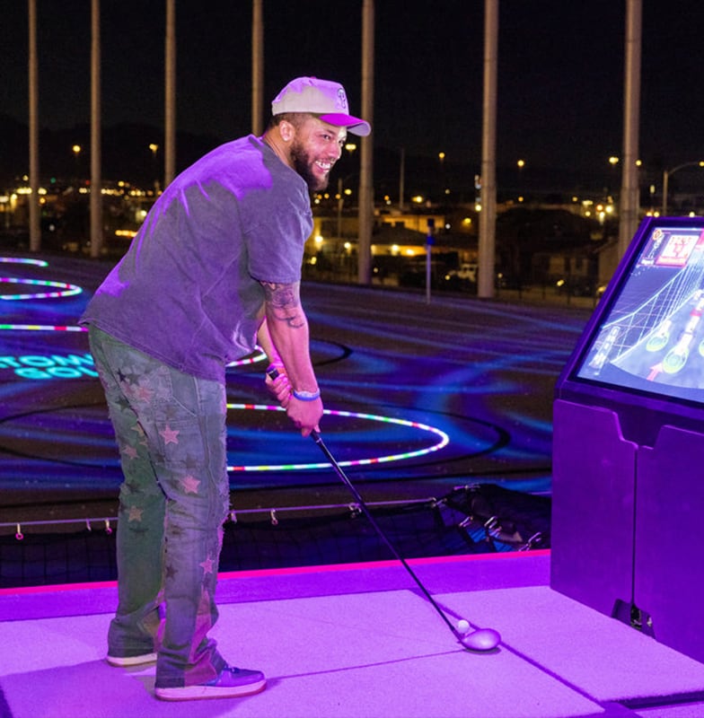 Man smiles while playing golf at a driving range with colorful lights and targets in the background at night.