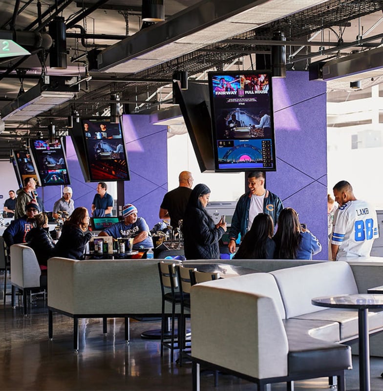 People gather in a modern sports bar with large TVs, watching games and socializing at tables and sofas.