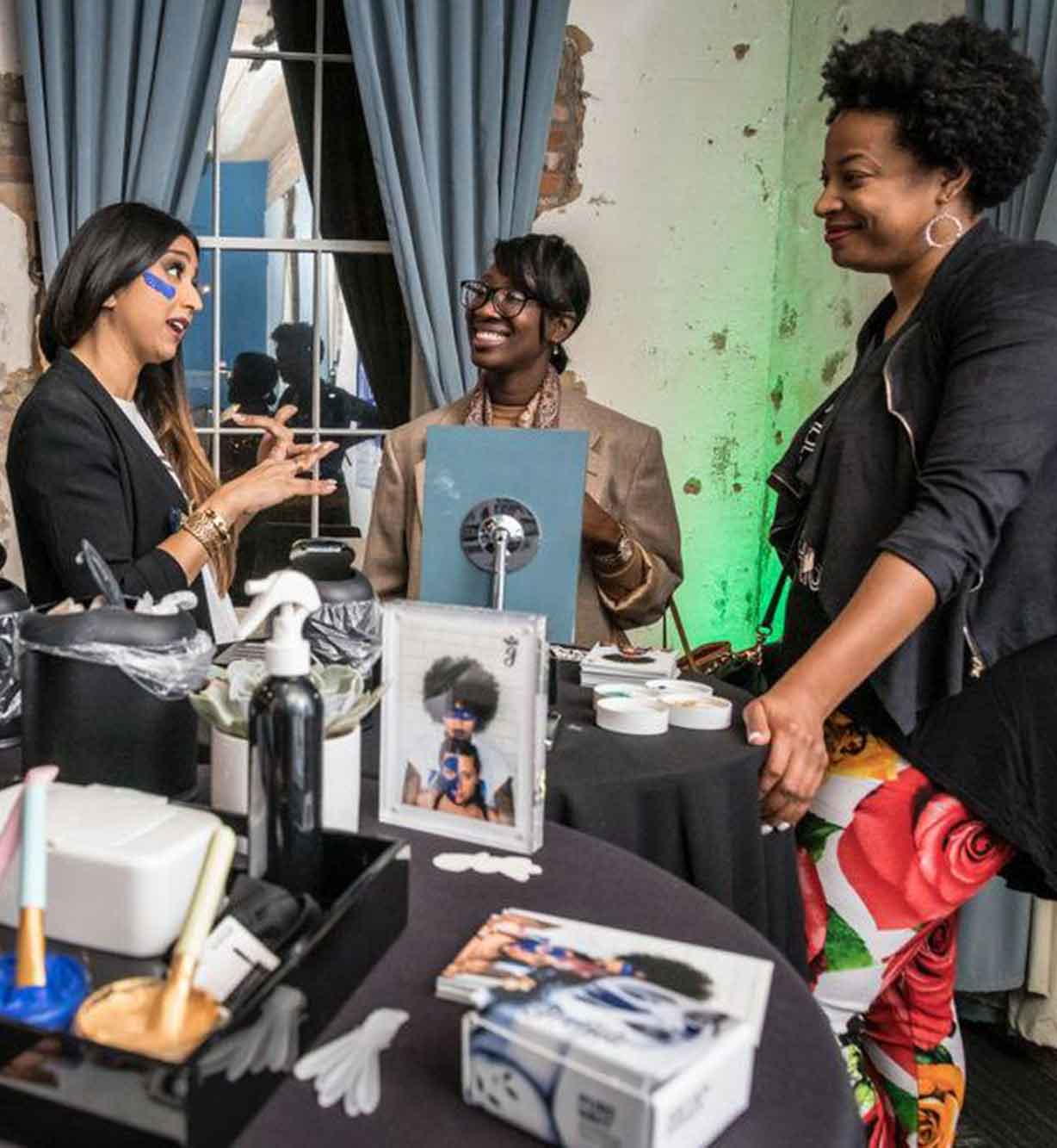Three women smiling and talking at a beauty product display table in a stylish indoor setting.
