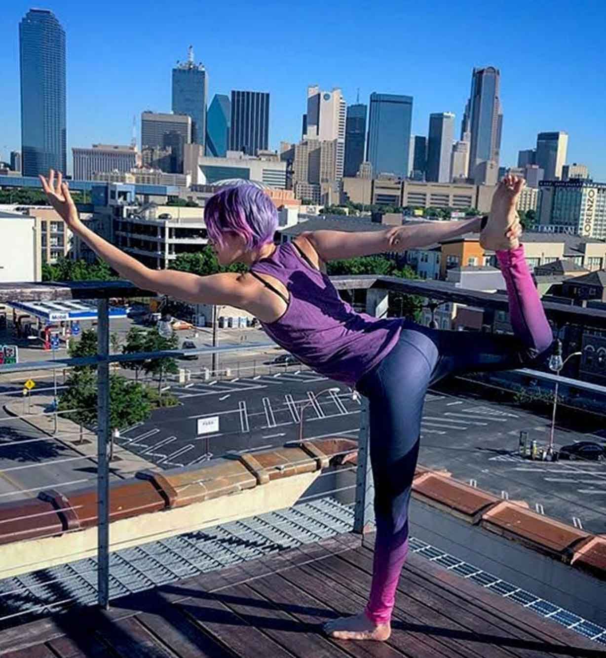 Person with purple hair does yoga pose on a rooftop with a city skyline in the background.