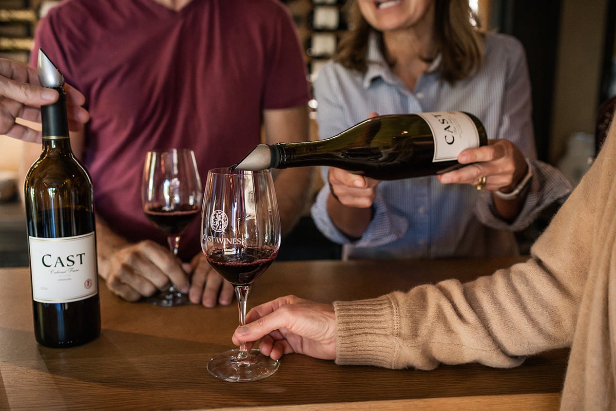 A person pours red wine into a glass at a bar, with two people smiling in the background.