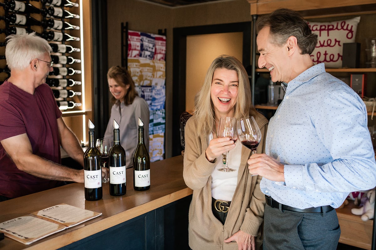 Four adults tasting wine and laughing together inside a cozy winery tasting room, bottles on the counter.