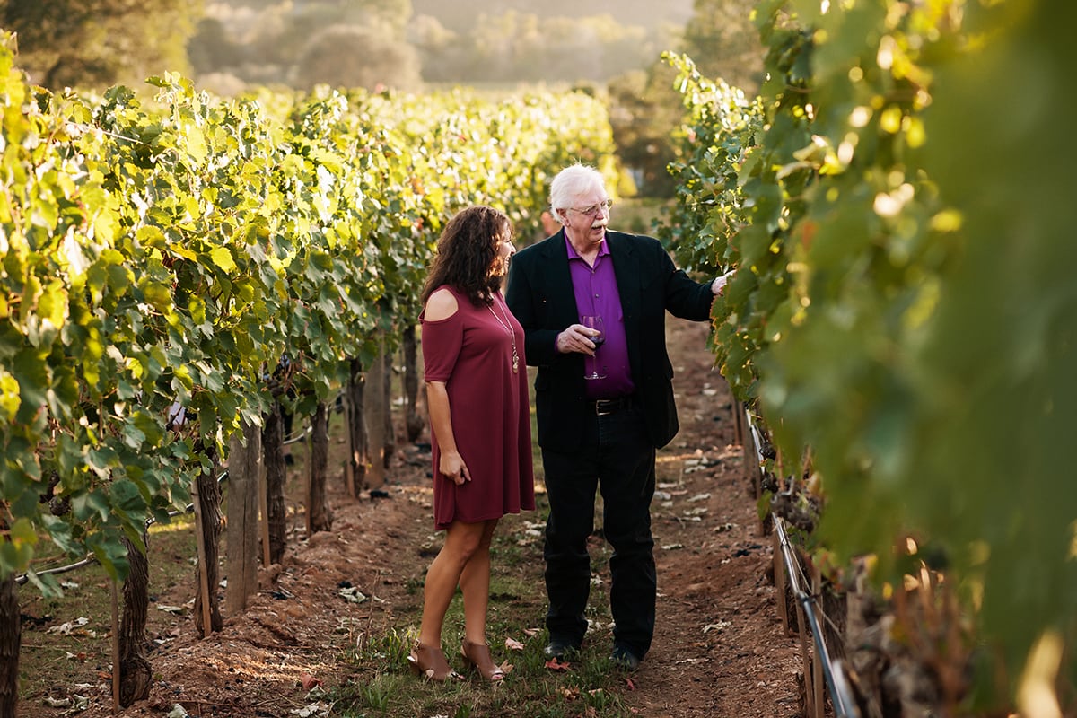 A man and woman walk through a sunlit vineyard, examining grapevines together.