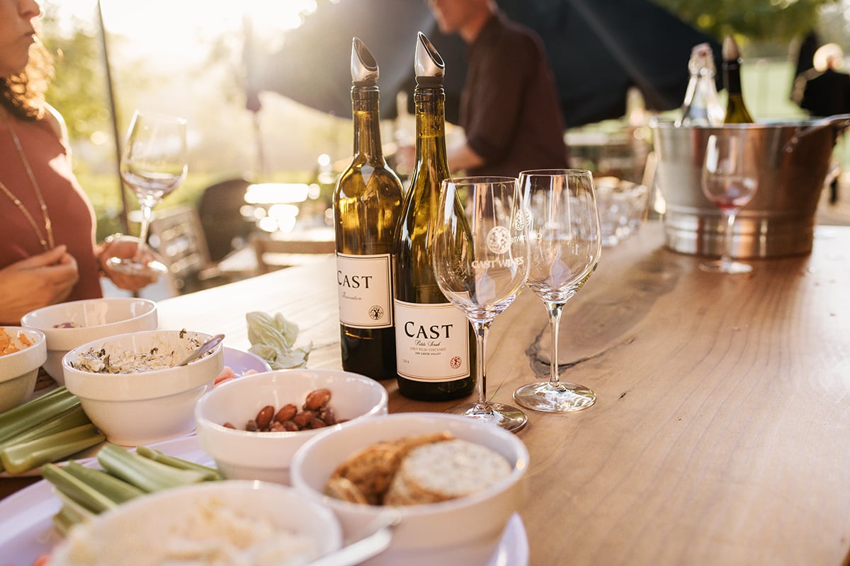 Two wine bottles and glasses on an outdoor table with snacks and sunlight in the background.