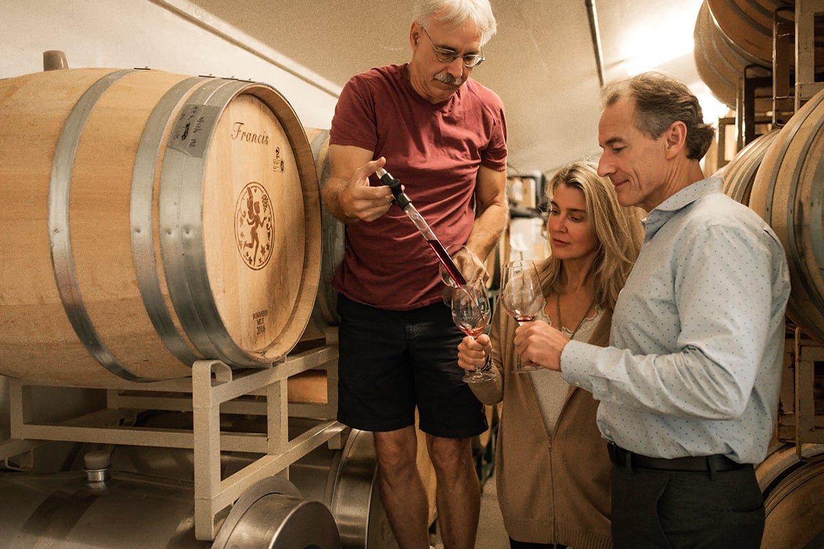 A man pours wine from a barrel into glasses held by a woman and another man in a cellar.