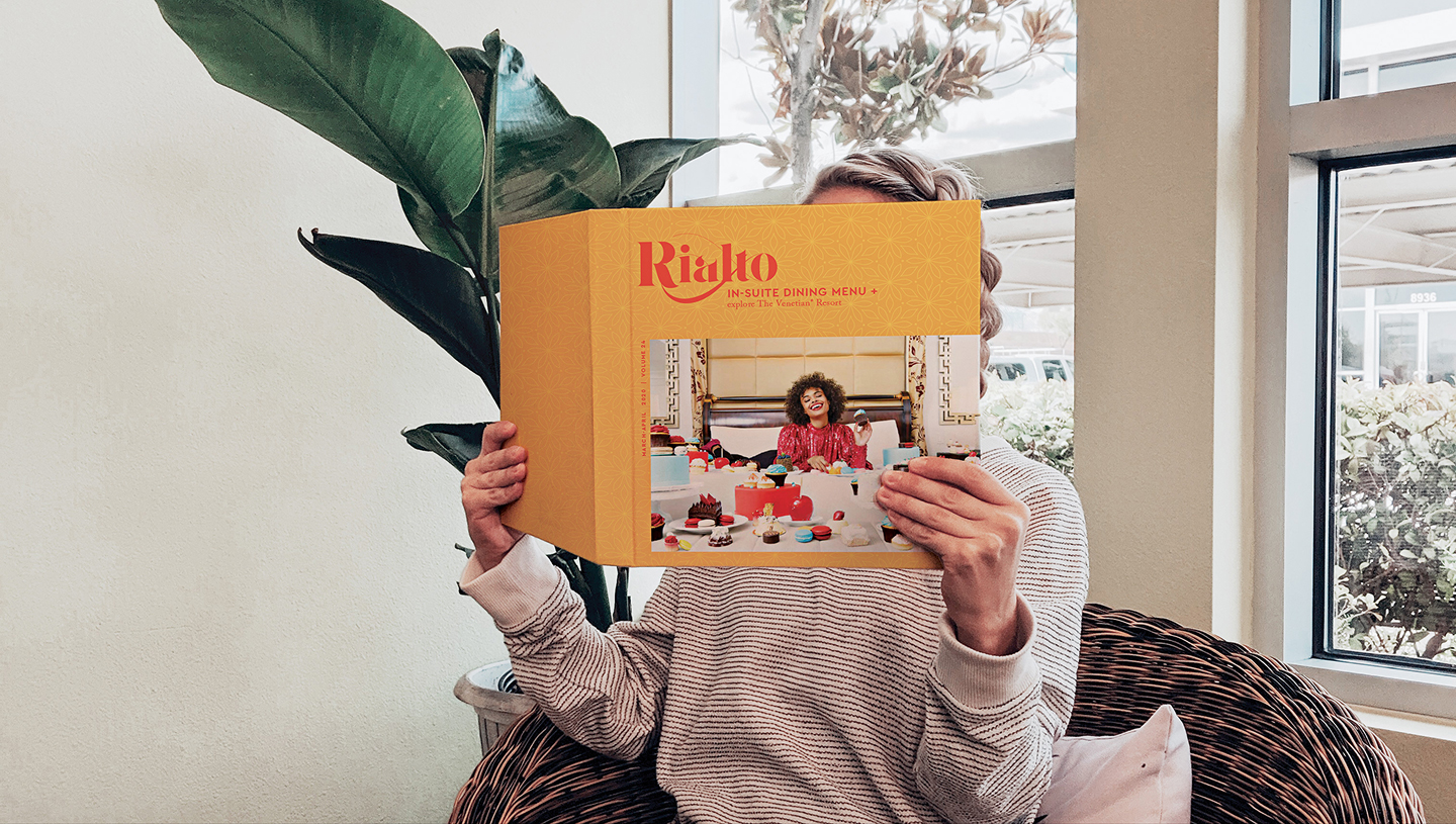 Person sitting in a chair, holding and reading a large yellow Rialto menu that covers their face.