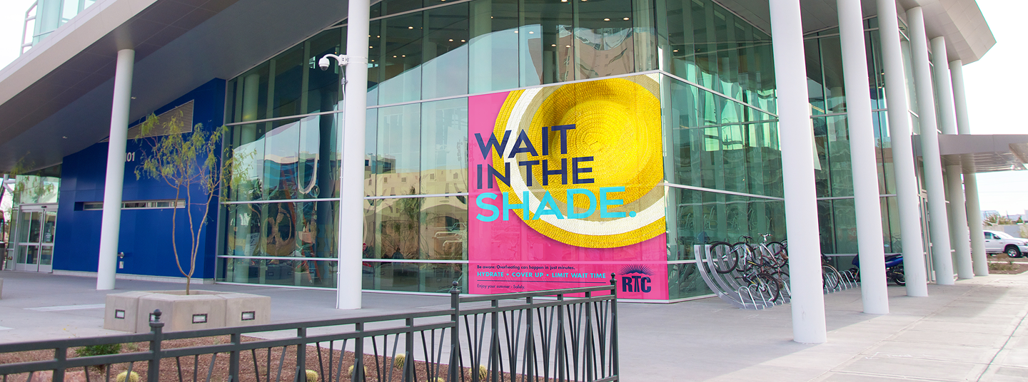 A modern glass building displays a large colorful poster reading WAIT IN THE SHADE near its entrance.