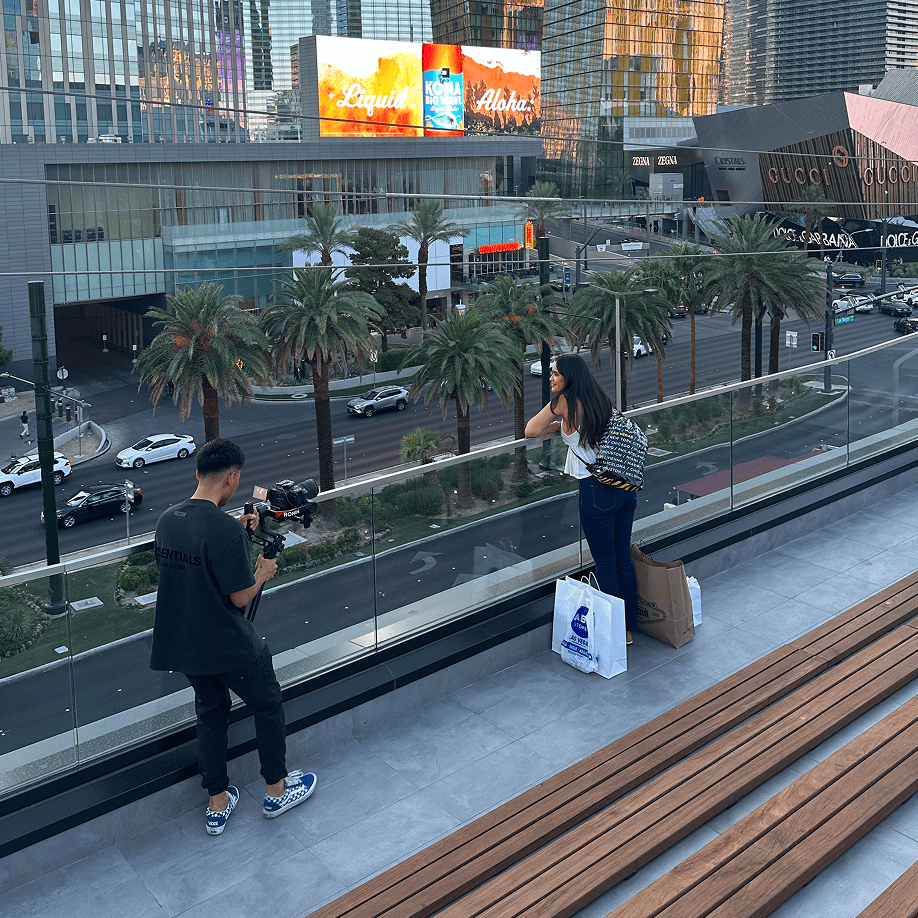 A man films a woman posing by a railing on a city street with palm trees and shops in the background.