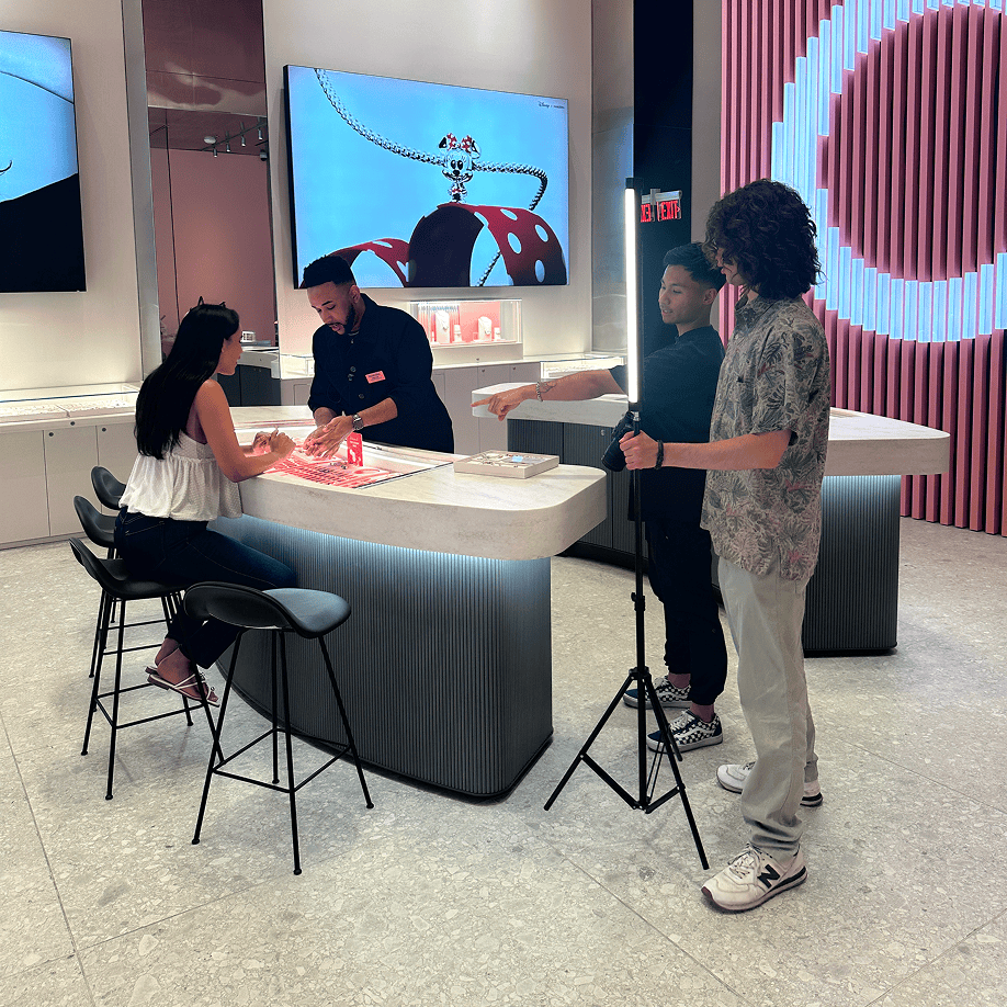 Two people sit at a store counter with a staff member, while two others film them with a camera and tripod.