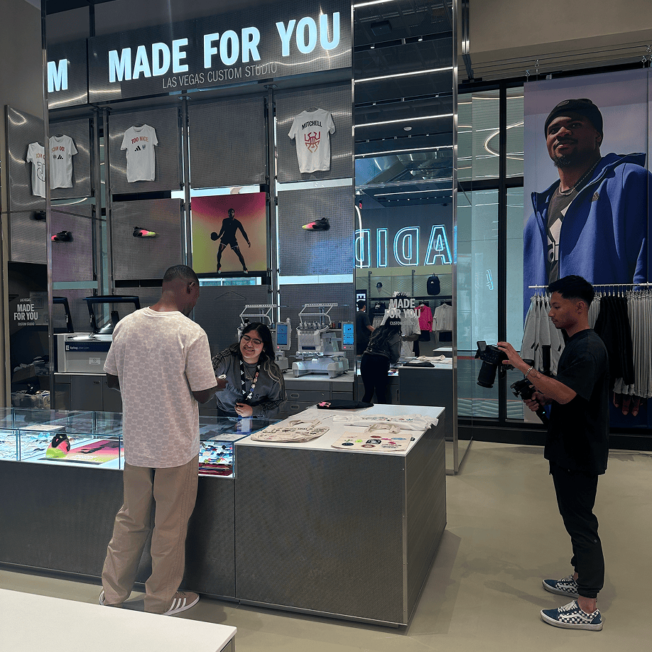Three people interact inside a modern clothing store with custom t-shirts and large display screens.