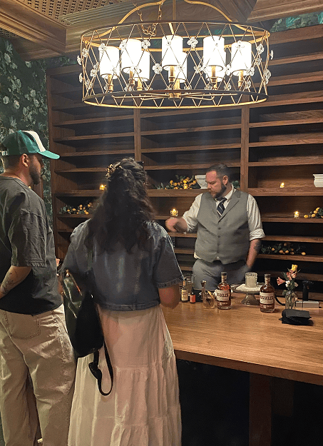 Three people at a bar with a bartender serving drinks under a decorative chandelier and wooden shelves.