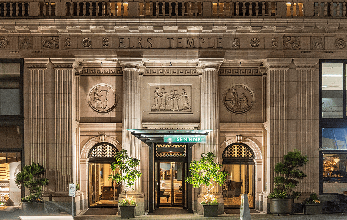 Night view of a grand, ornate building entrance labeled ELKS TEMPLE with “Sentinel” sign and potted plants.