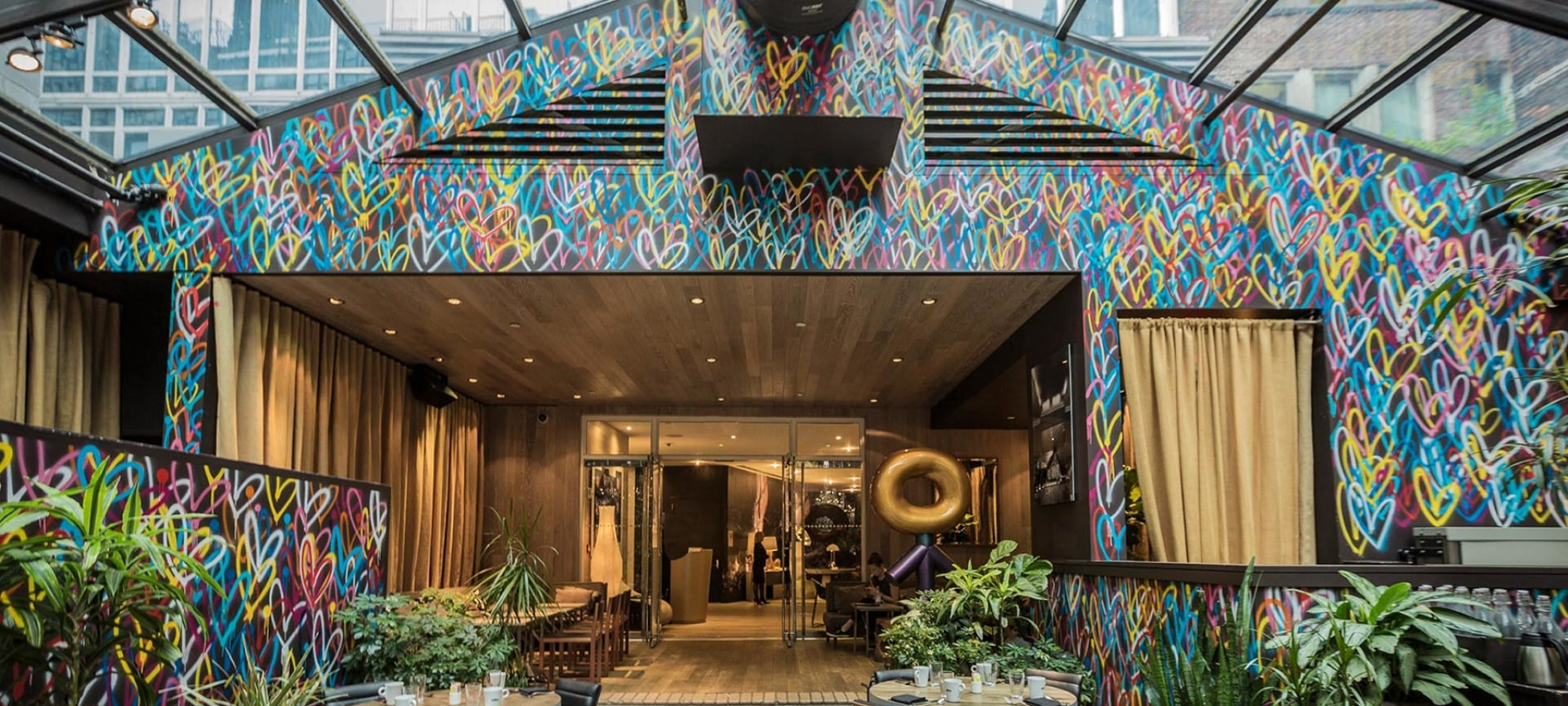 Colorful heart graffiti covers the exterior walls of a cafe with plants and a glass ceiling.