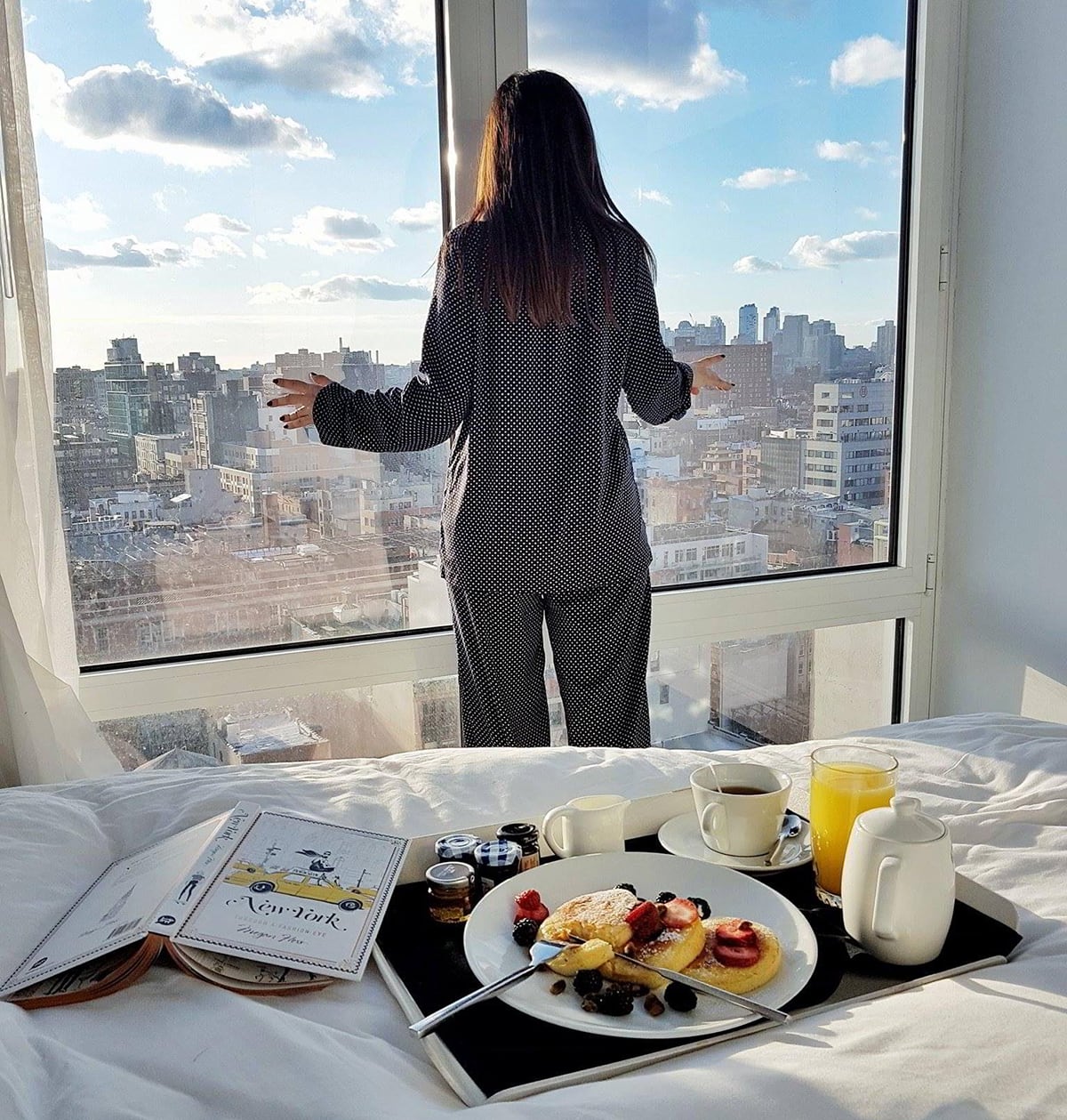 Woman in pajamas stands by a window overlooking a city, with breakfast and a book on the bed in front.