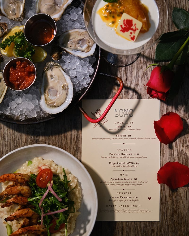 A Valentines Day dinner with oysters, shrimp risotto, a cocktail, red roses, and a printed menu on a table.