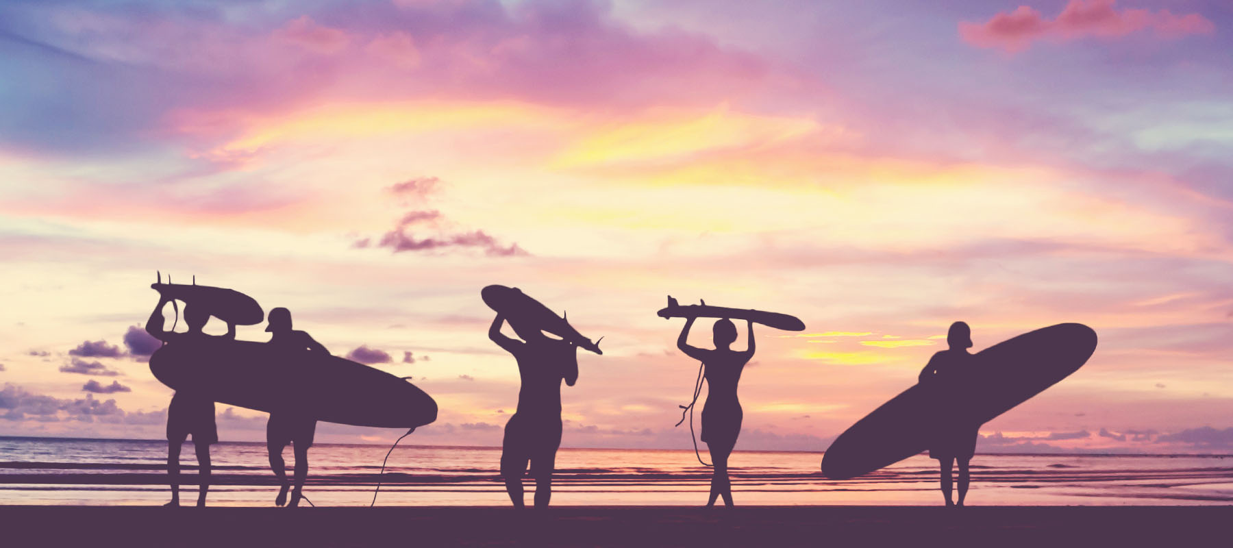 Five surfers hold surfboards on the beach at sunset, silhouetted against a colorful sky.