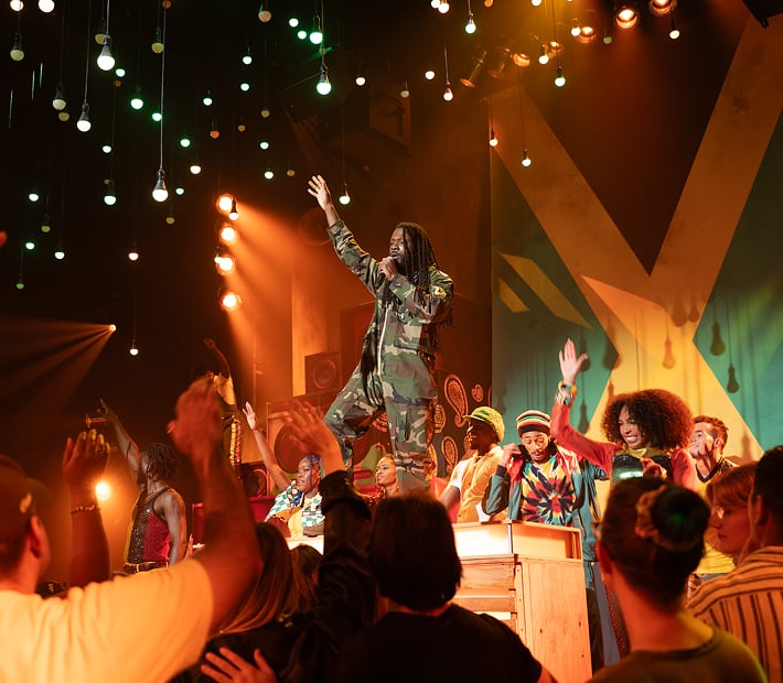 Performer in camouflage sings on stage, surrounded by energetic dancers and cheering crowd under bright lights.