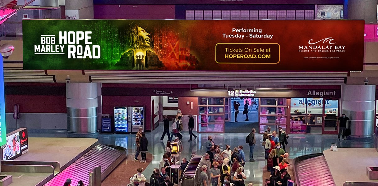 Large Bob Marley Hope Road show billboard above airport baggage claim; travelers gather below.