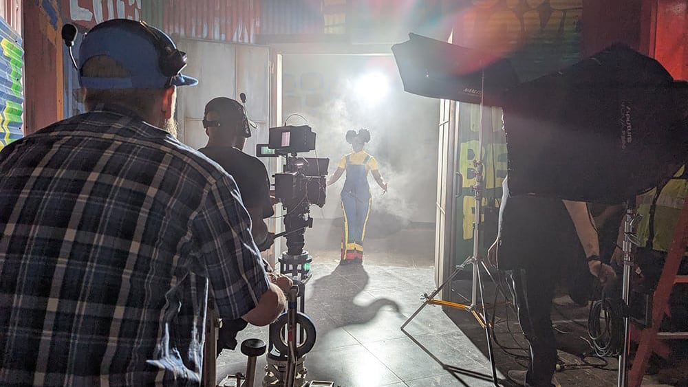 Film crew shoots a woman in costume walking through a smoky doorway with bright lights behind her.